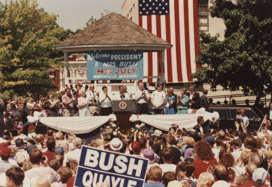 A crowd of people watches as President George H. W. Bush speaks at the 1991 Fourth of July parade in Marshfield. Bush speaks on a stage in front of the courthouse which is draped with a large American flag. Sign on gazebo reads: "Welcome President & Mrs. Bush 4th of July Marshfield, Missouri"