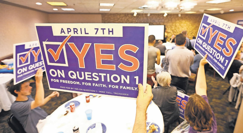 People sitting at a table in a restaurant hold up signs reading "April 7th. Yes on Question 1. For Freedom. For Faith. For Family. Paid for by Yes on Question 1 - Mike Tull, Treasurer."