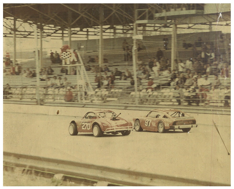 Two red race cars driven by Roger Arnhart and Jerry McCall respectively approach the checkered flag at the Fairgrounds Speedway in Springfield, Missouri. A few dozen people are seated in the grandstand.