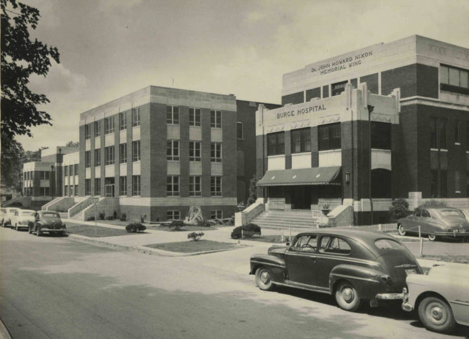 A black and white photograph of Burge Hospital in Springfield. Burge is a 3-4 story building. Cars are parked on the street in front of the hospital's entrance.