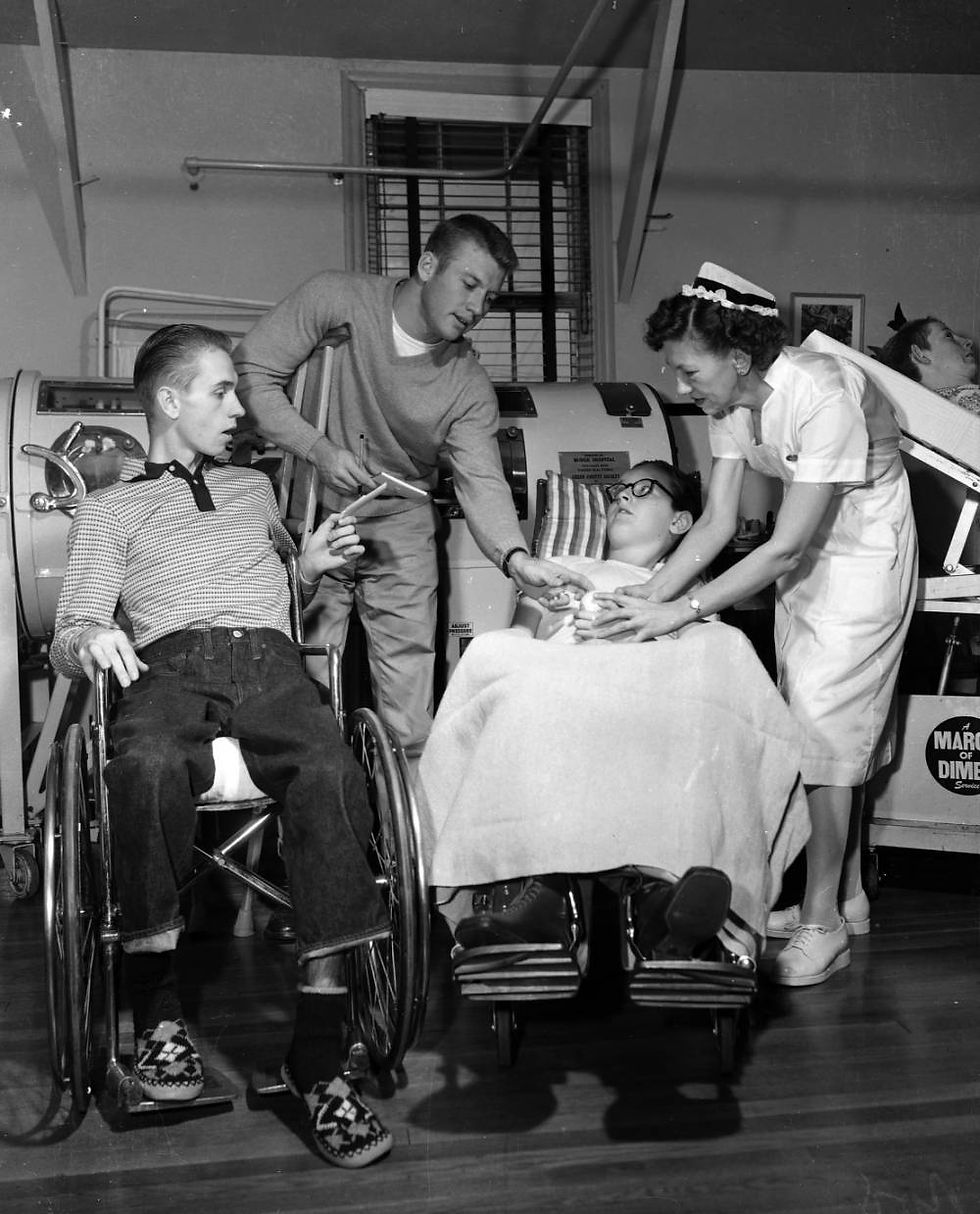 A black and white photograph of Mickey Mantle, leaning on a crutch, handing an autographed baseball to Gwinn Hinkle, a young boy who is in a wheelchair, in the polio ward of Burge Hospital. Another boy in a wheelchair, Clark Spears, looks on from the left, while an unidentified nurse helps Gwinn to grasp the baseball.