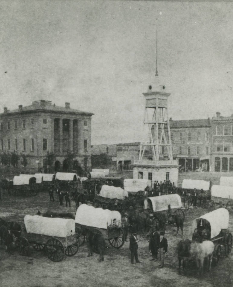 A black and white photograph of Springfield's public square in the 1880s. Horses and wagons fill the square. A water tower is in the center of the square. The Greene County Courthouse stands at left.