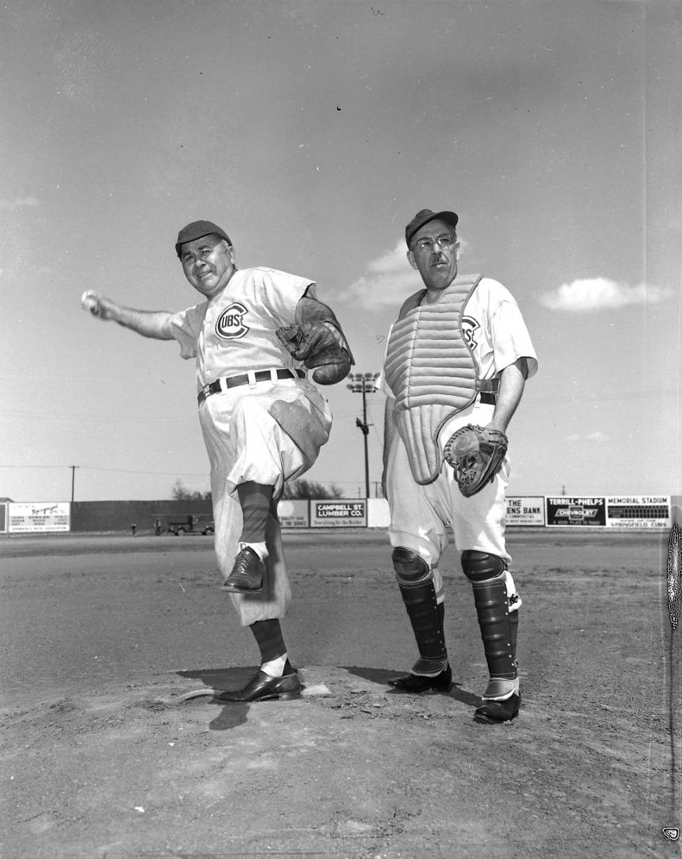 A black and white photograph of C. C. Williford and Otis L. Barbarick in Springfield Cubs uniforms, standing on the mound at Memorial Stadium. Williford, a heavyset man, is rearing back to pitch. Barbarick wears glasses and catcher's gear.