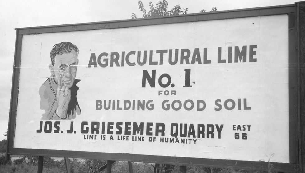 A black and white photograph of a sign for Griesemer Quarry. A posterized illustration of a man lifting up one finger, indicating that the quarry's product is "number one," is on the left of the sign. Text on the sign reads: "Agricultural Lime. No. 1 for Building Good Soil. Jos. J. Griesemer Quarry. East 66. Lime is a Life Line of Humanity."