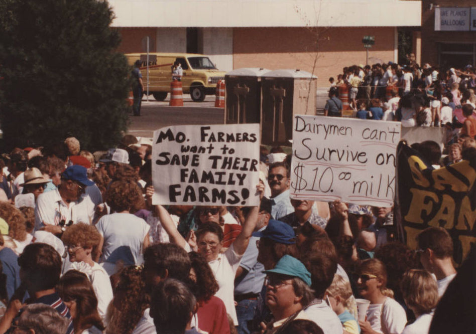 Protestors hold up signs in the crowd at Marshfield's 1991 Fourth of July parade. Signs read: "Mo. Farmers want to save their family farms!" and "Dairymen can't survive on $10.00 milk"