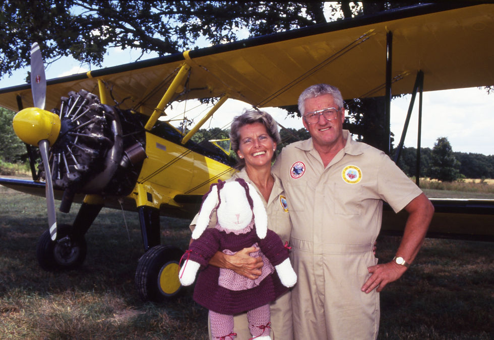 Jody and Dave Corsaut, both wearing jumpsuits, stand in front a yellow Stearman biplane. Jody is holding a large knitted rabbit.