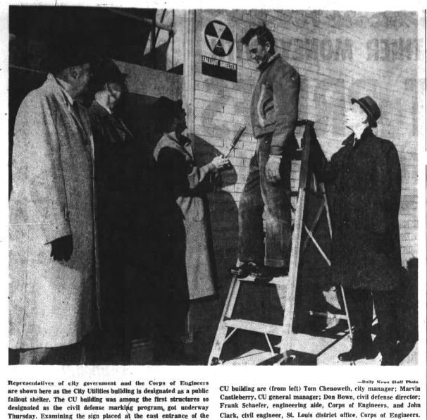 A clipping from the December 14, 1962 issue of the Springfield Daily News. Several officials stand in front of the eastern entrance of the City Utilities building, admiring a newly-placed "Fallout Shelter" decal designating the building as a community fallout shelter. Text reads:
"Representatives of city government and the Corps of Engineers are shown here as the City Utilities building is designated as a public fallout shelter. The CU building was among the first structures so designated as the civil defense marking program got underway Thursday. Examining the sign placed at the east entrance of the CU building are (from left) Tom Chenoweth, city manager; Marvin Castleberry, CU general manager; Don Bown, civil defense director; Frank Schaefer, engineering aide, Corps of Engineers, and John Clark, civil engineer, St. Louis district office, Corps of Engineers."