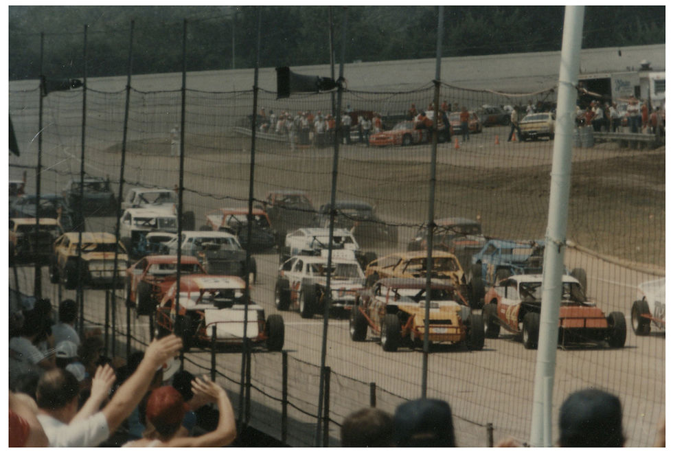 Racers in cars of various colors complete a pace lap before the start of a race at the Fairgrounds Speedway. Spectators can be seen in the stands and in the infield.