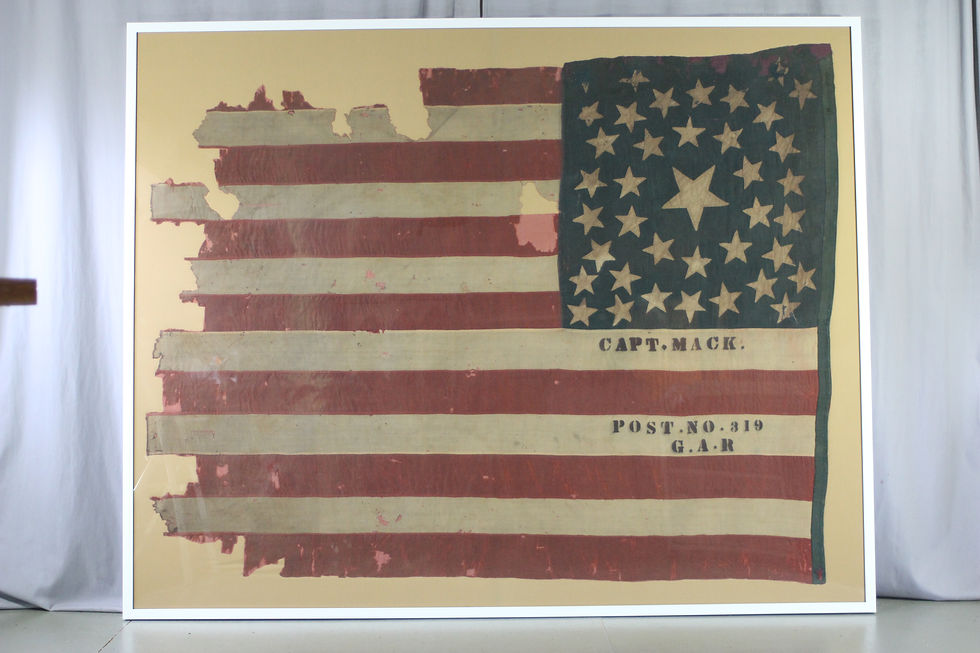 A large, tattered American flag. The stars on the flag are arranged in a circle. Text on two of the white stripes reads: "Capt. Mack. Post No. 319 G.A.R."