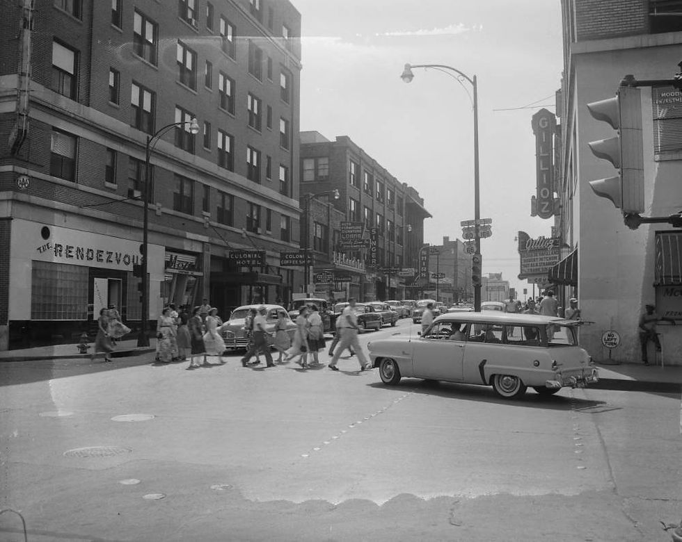 A black and white photograph of pedestrians crossing St. Louis Street near Jefferson Avenue in Downtown Springfield. The pedestrians are dressed in mid-1950s stylers. A vehicle drives near the pedestrians.