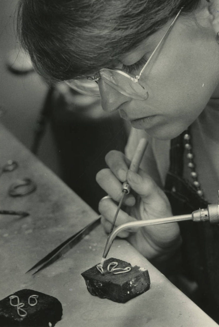 A black and white photograph of Elaine Cearlock, a young woman, using a blowtorch and a pick to shape pieces of metal jewelry.