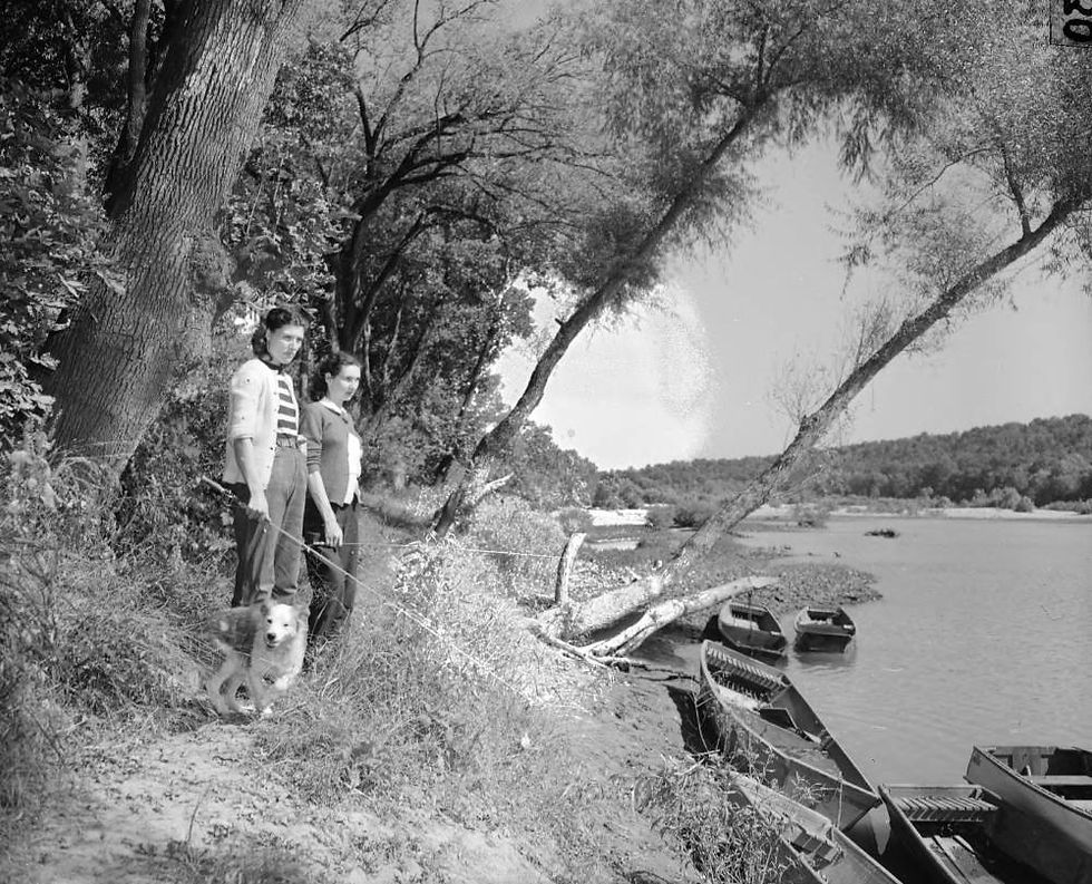 Two young girls holding fishing poles stand with a dog on the wooded banks of the White River. Several boats are docked nearby.