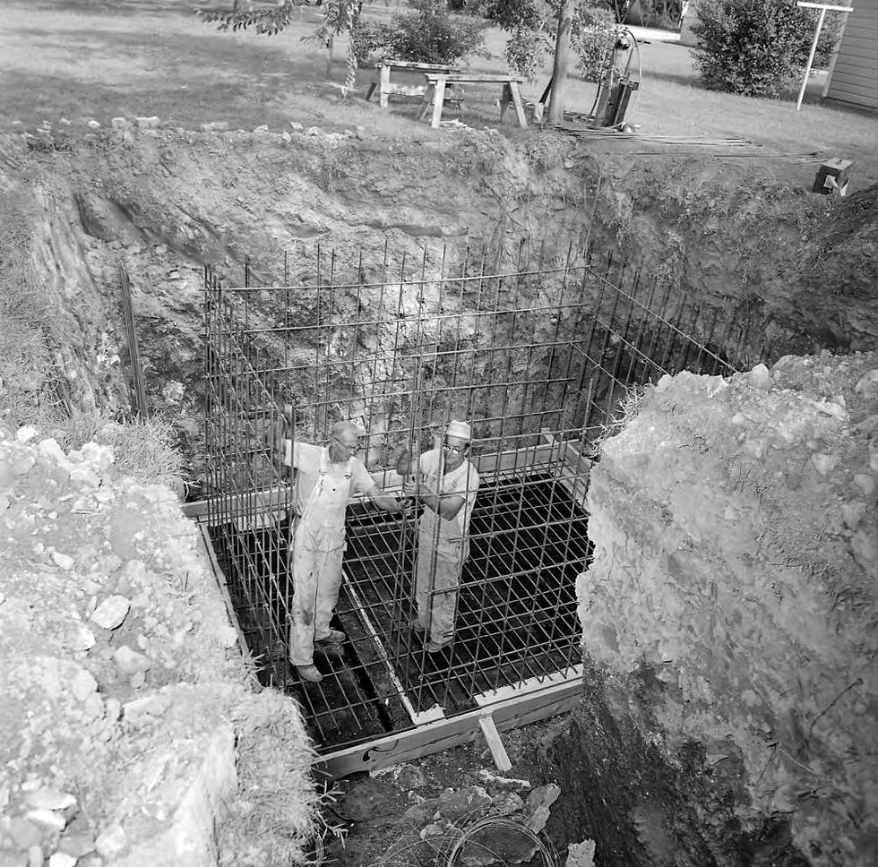 Two men, James R. Jeffires and James L. Maynard, work on installing an underground fallout shelter in the yard of Mr. and Mrs. Donald L. Price. The two men stand in a deep hole in the ground, laying the supports for what will become the walls of the fallout shelter.