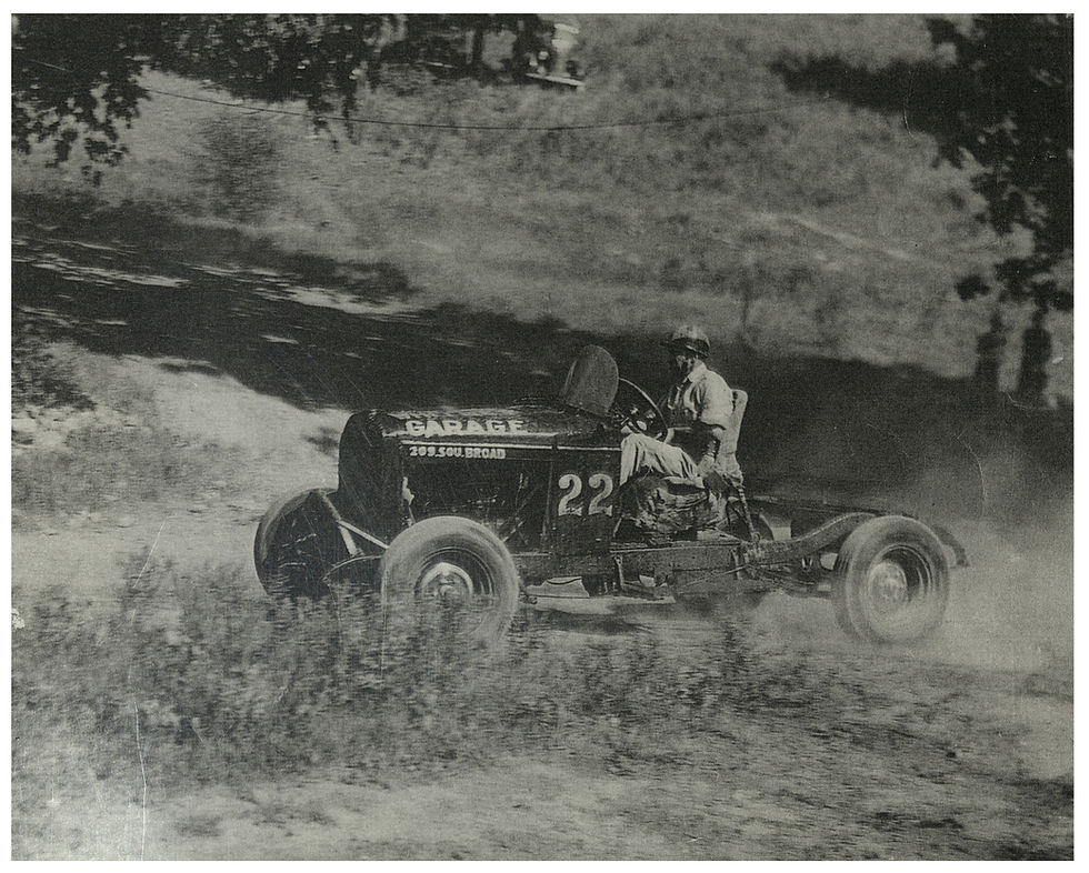 A black and white photograph of racer Si Whitney driving a jalopy in a race at the Perry Smith Farm. He drives on a dirt track in a rural setting.