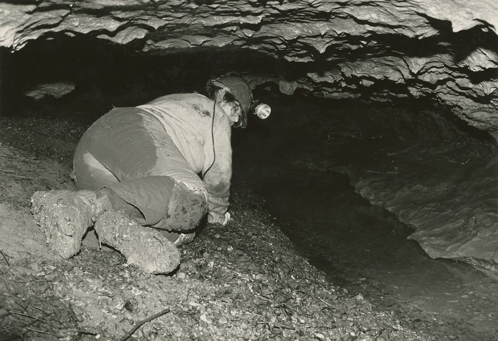 A black and white photograph of Jo Schaper in coveralls, boots, and a helmet with a headlamp crawling through a low passage in Fantastic Caverns. At right is a pool of water.
