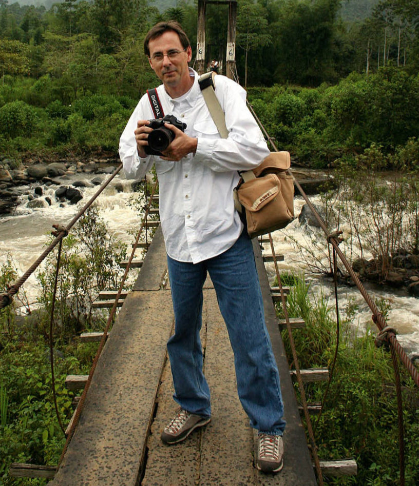Bob Linder stands on a suspension bridge in Ecuador, holding a digital camera. A river runs in the background beneath the bridge.