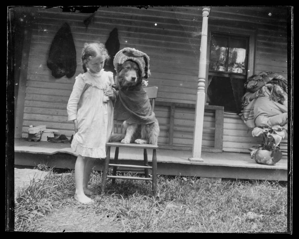 A black and white antique photograph of a young barefoot girl posing with a dressed-up dog in front of a porch.