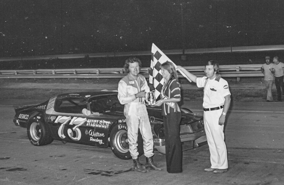Dave Watson receives the trophy for the 1976 Missouri State Asphalt Championship from an unidentified woman while flagman Bill Crooks holds the checkered flag.