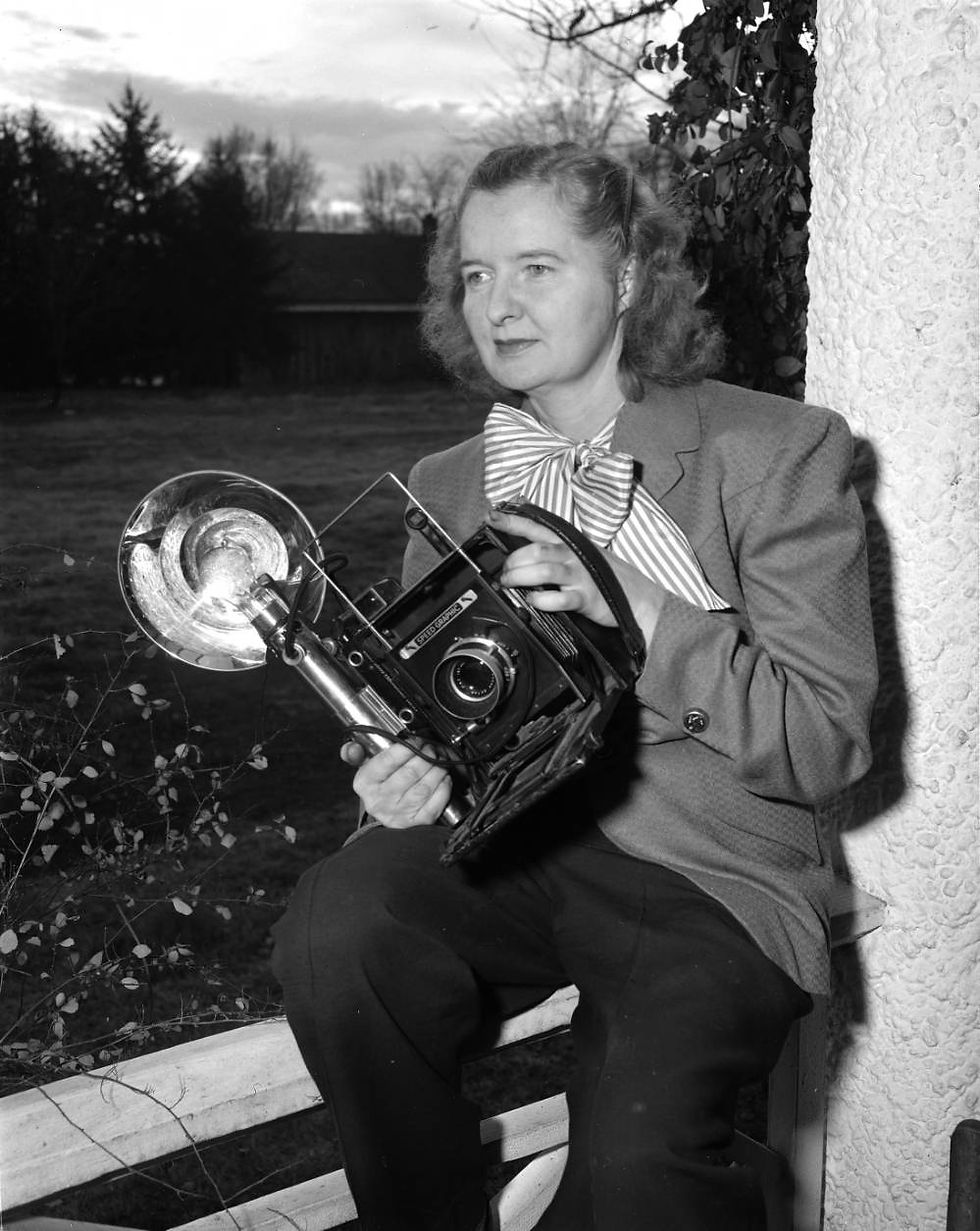 Betty Love poses with her camera. Love is seated on a railing; a yard is in the background. She holds a camera with a large flash bulb.