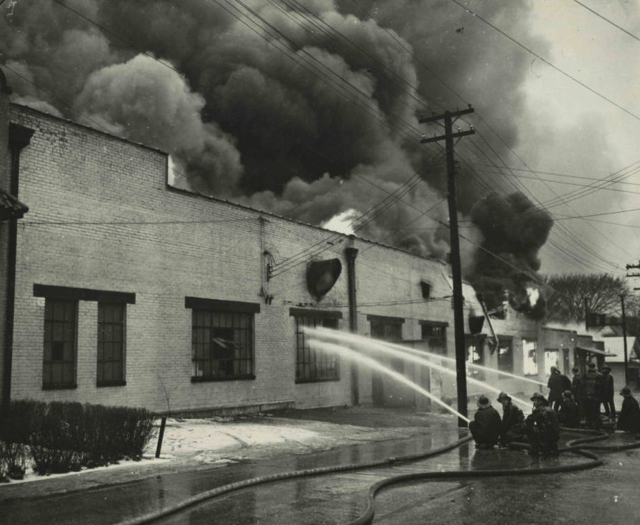 Firefighters working to extinguish a fire at the Springfield Newspapers, Inc. offices on March 27, 1947. A large cloud of dark smoke rises in the background behind the building.