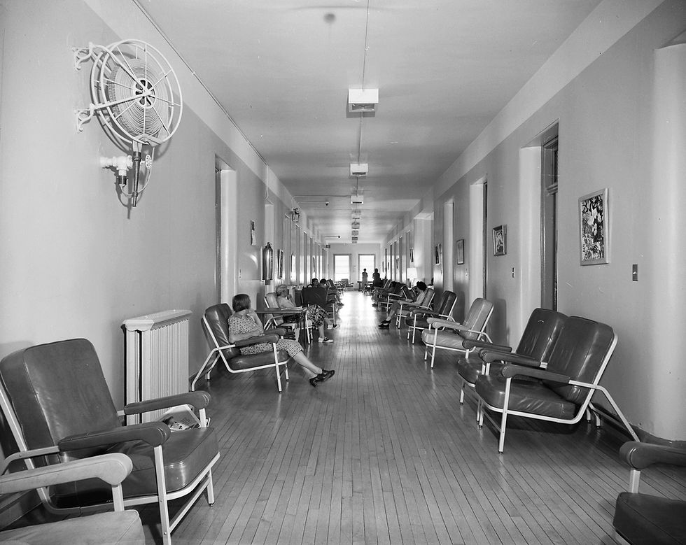 Black and white photograph of a hallway at State Hospital No. 3. Chairs line both sides of the hallway; patients sit in a few of the chairs.