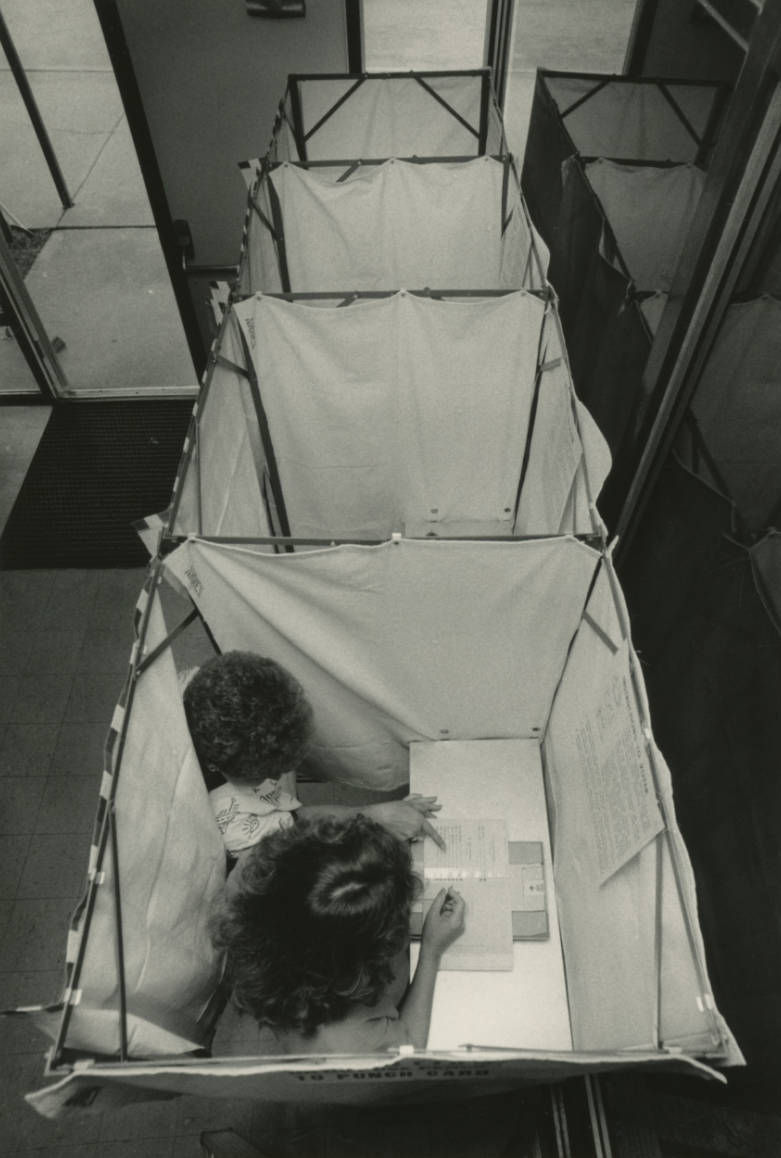 Black and white photograph of an election judge showing a woman how to fill out a ballot inside of a voting booth. 