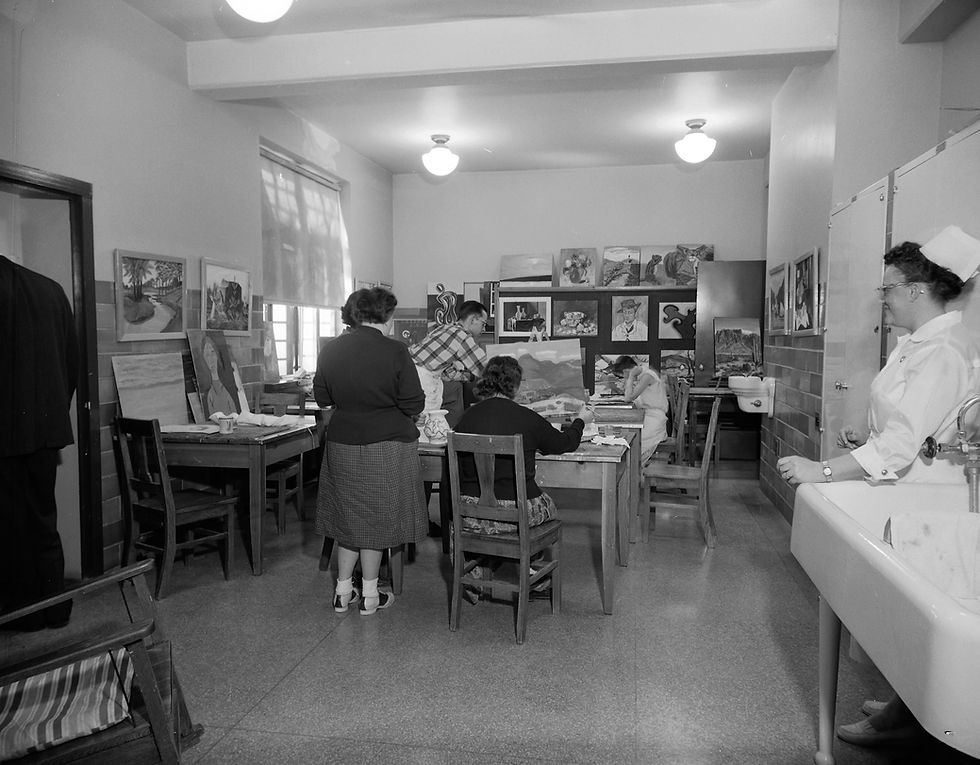 Patients work in the art therapy department's studio at State Hospital No. 3. Patients sit and stand around a table; some painting. A nurse supervises.