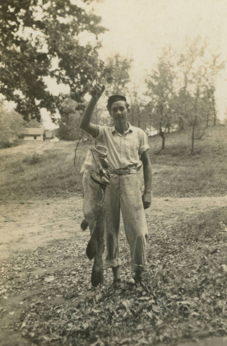 A sepia-tone photograph of a man standing outdoors, holding a large bass by a string.