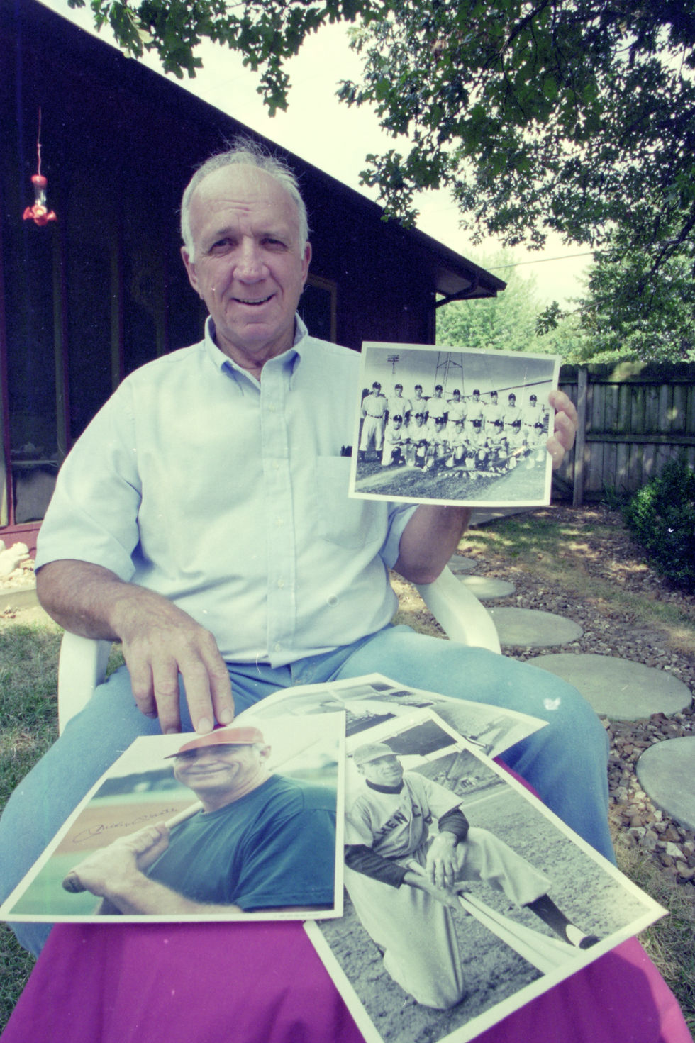 Jack Hasten holds up a photograph of a baseball team. Hasten is an older man with wispy white hair. He is seated in a yard. More baseball photographs are displayed on a small table in front of him.