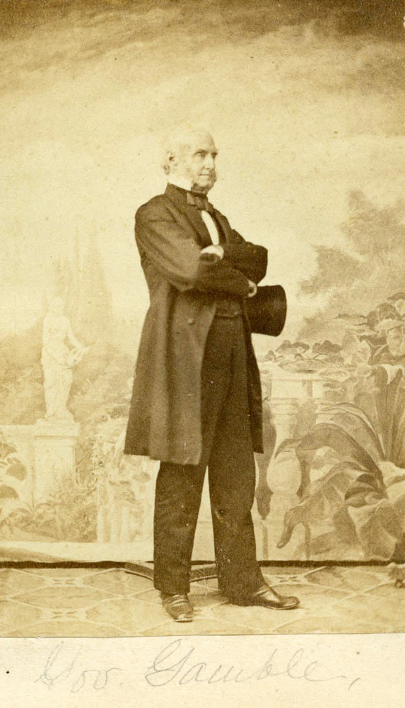 A full length portrait of Governor Hamilton Gamble. Gamble is an older white man. He is bald and has short facial hair. He holds a hat and is wearing a suit.