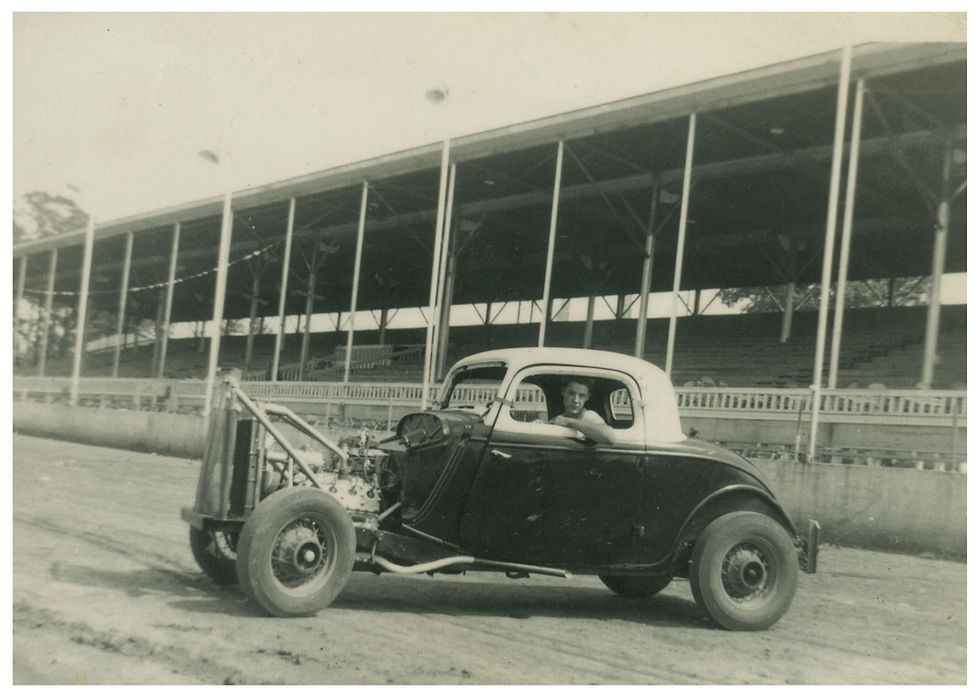A black and white photograph of racer Bill Chandler in his race car. The car is parked in front of a grandstand and is on a dirt track at the Fairgrounds Speedway.