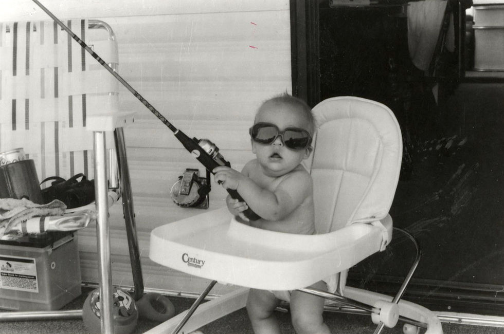 A black and white photograph of a diaper-clad infant sitting in a highchair, wearing sunglasses, and holding a fishing pole.