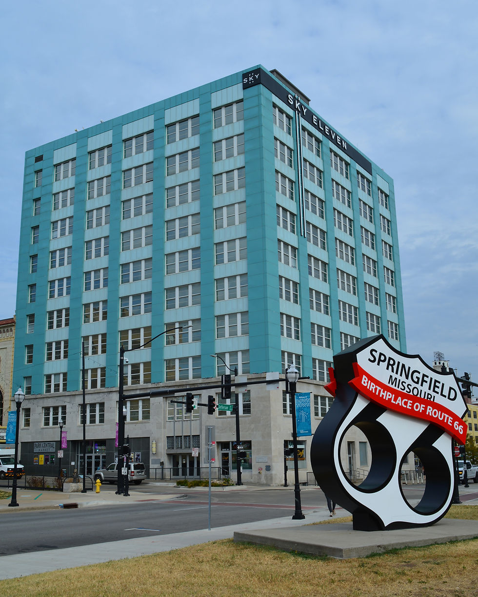 A contemporary photograph of Sky Eleven (once the Woodruff Building) and the nearby sculpture of the Route 66 highway shield. Text on the sculpture reads "Springfield Missouri: Birthplace of Route 66." Sky Eleven's facade is a robin's egg blue.
