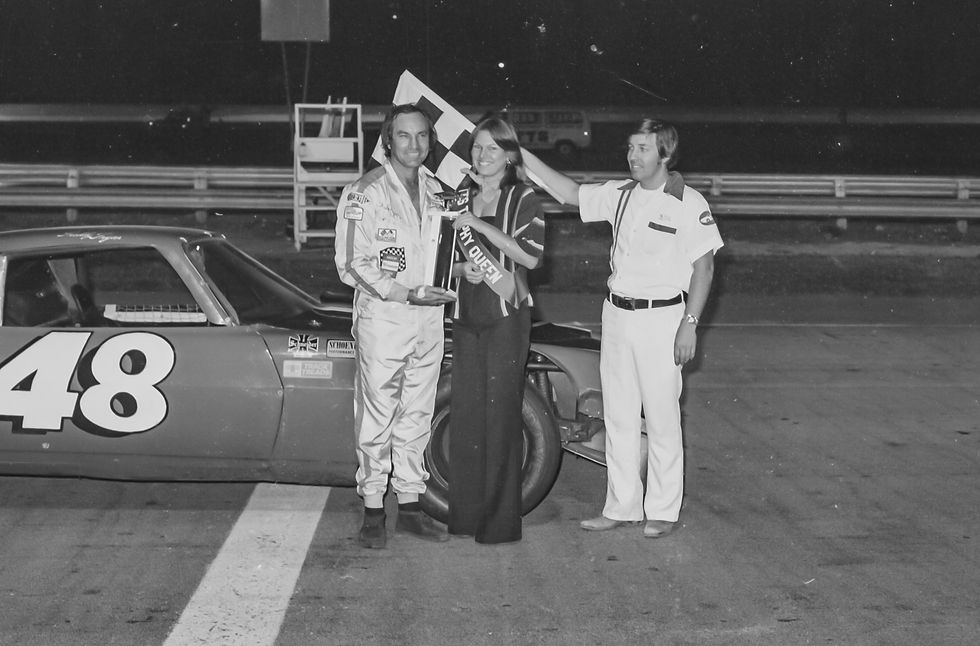 Freddy Fryar poses with a trophy in victory lane after winning the trophy dash of the 1976 Missouri State Asphalt Championship. An unidentified woman presents Fryar with the trophy while flagman Bill Crooks holds the checkered flag.