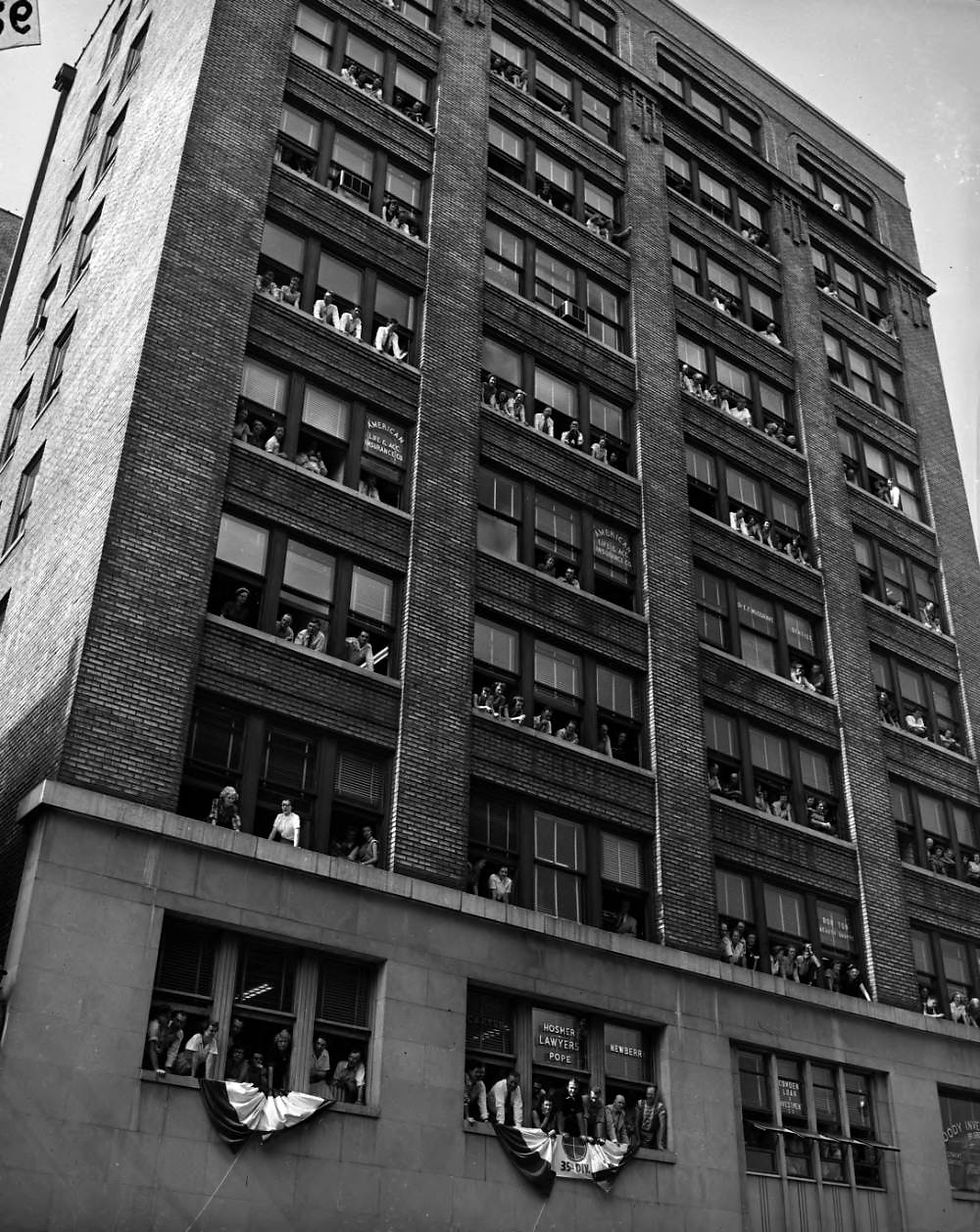 A black and white photograph of Woodruff Building employees leaning out of the building's windows. The building has ten stories and a brick facade.