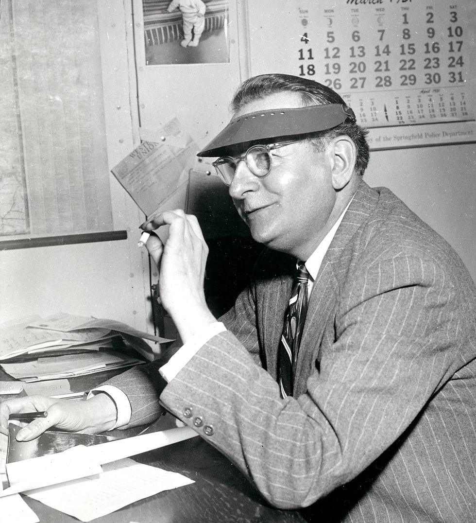 George Olds, former editor-in-chief for Springfield Newspapers, Inc., seated at a desk and smoking a cigarette. Olds wears eyeglasses, a visor, and a suit and tie.