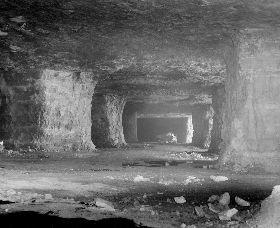 A black and white photograph of the interior of Griesemer Quarry, now known as the Springfield Underground. Large limestone "pillars" have been left in place at regular intervals, creating underground "rooms." A truck sits within the dark entrance of another room in the distance.