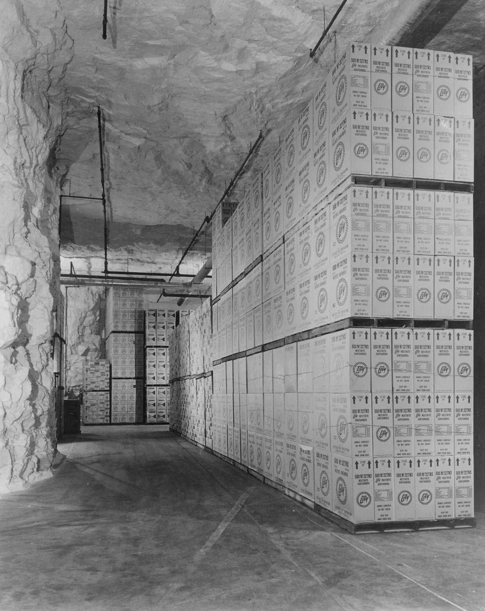A black and white photograph of an underground storage facility within Griesemer Quarry. Boxes of goods are stacked to the right of the frame on a concrete floor. The walls and ceiling of the room are roughly hewn limestone.