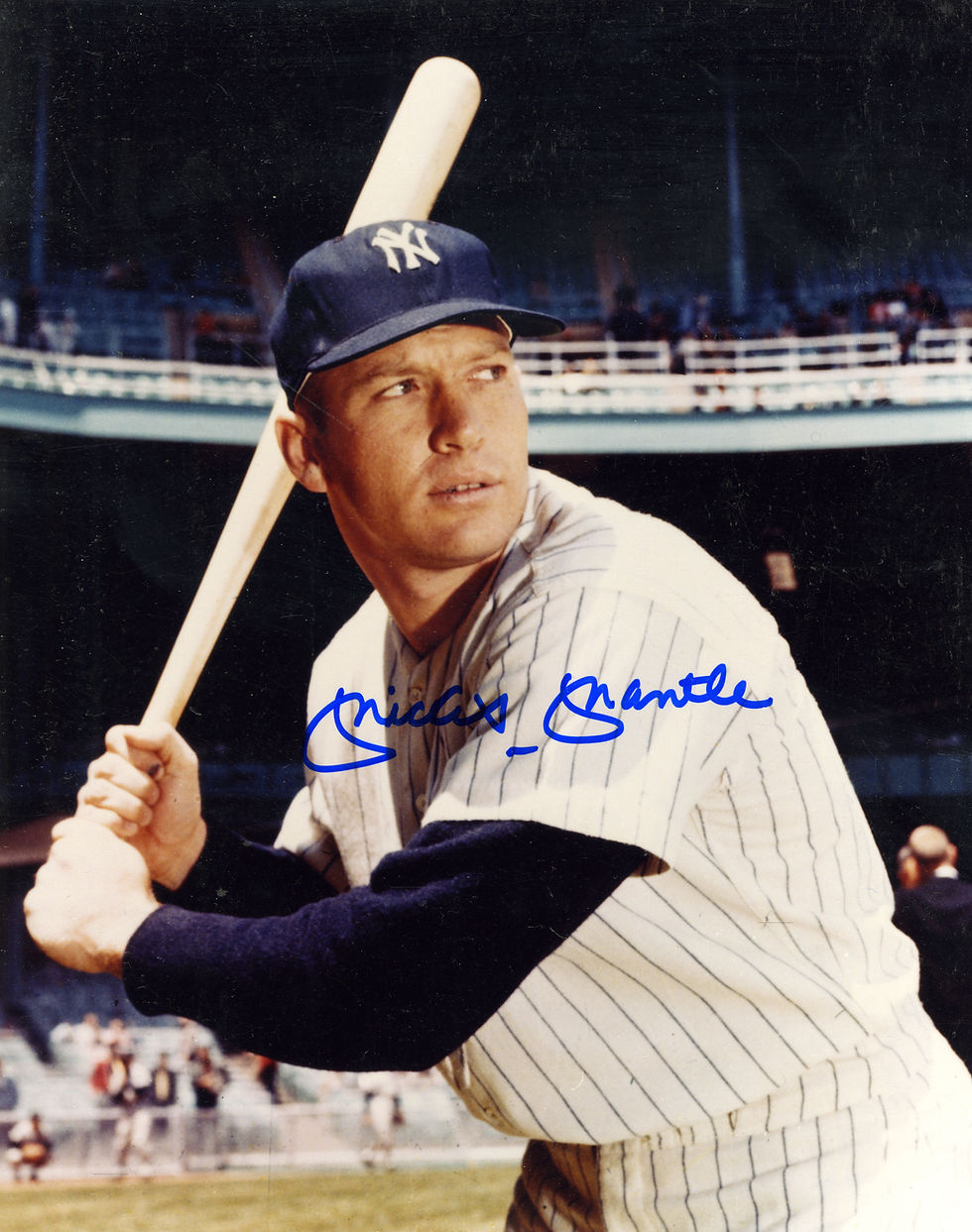 An autographed photo of Mickey Mantle standing on the mound at Yankee Stadium. Mantle is wearing a Yankees uniform and holding a bat. The photograph is signed with a blue marker.