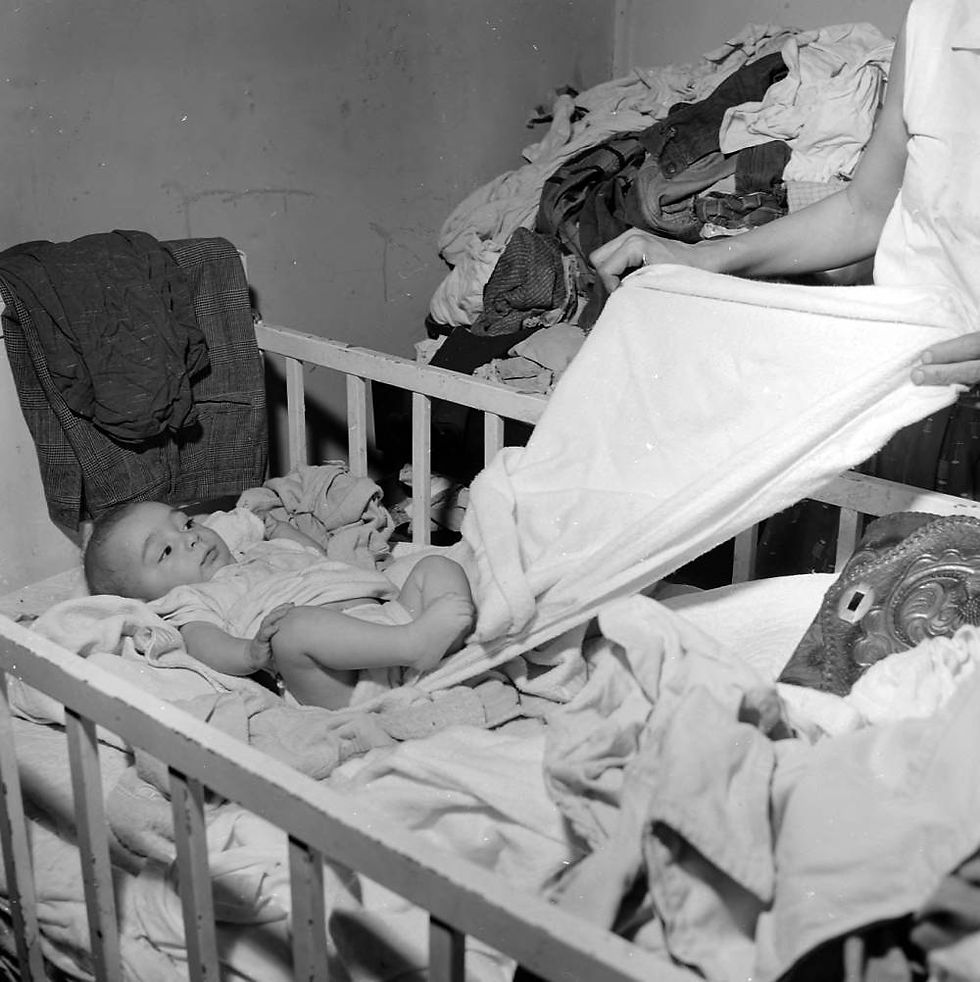 A black and white photograph of an infant laying in a cradle crowded with blankets and clothing. A woman pulls a blanket out from beneath the infant.