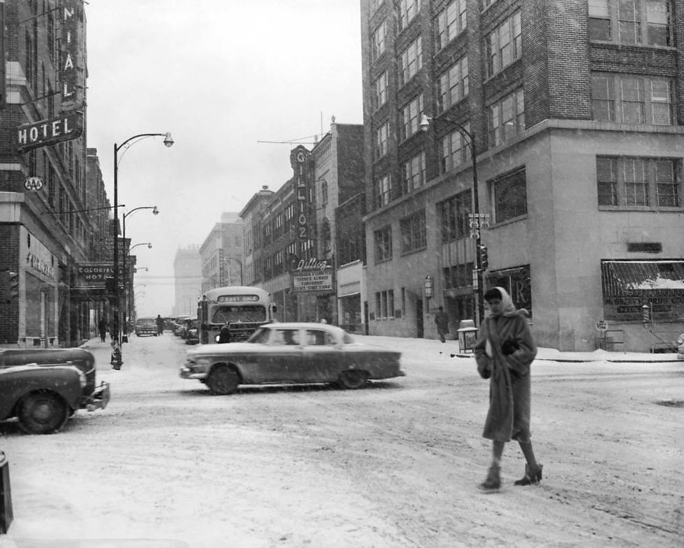 A black and white photograph of the intersection of Jefferson Avenue and St. Louis Street in Downtown Springfield during snowy weather. A woman crosses the street in the foreground; traffic passes by in the background.