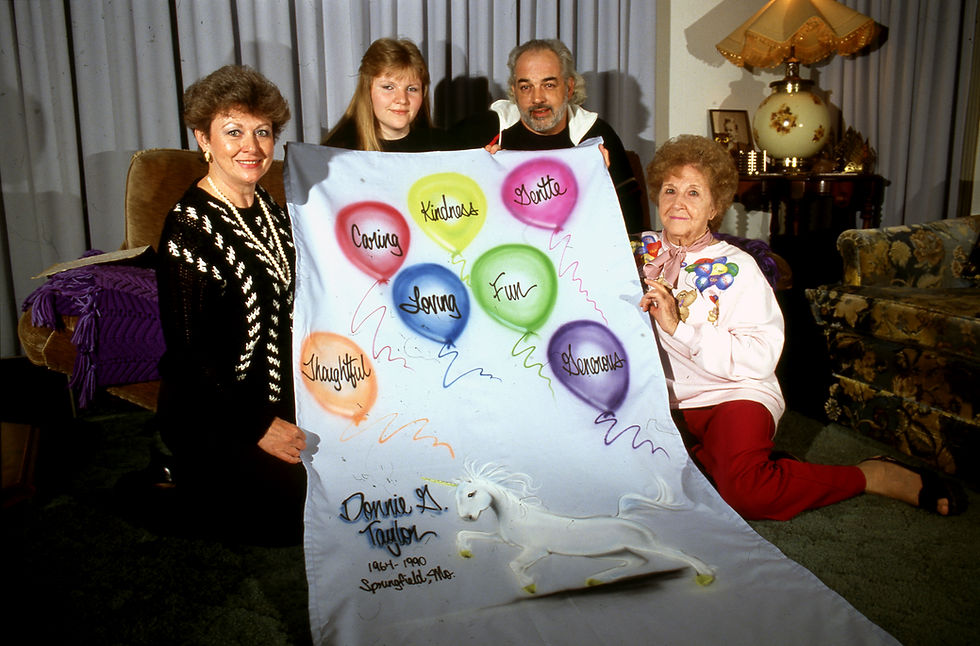 Loved ones of Donnie Taylor, who passed away from AIDS in 1990, gather around his block of the AIDS Memorial Quilt. From left, they are Pat Loveland, Joy Ransom, David Bridges, and Sylvia Cox. The quilt block features a unicorn and balloons with text inside of them. They read: "Thoughtful," "Caring," "Kindness," "Loving," "Fun," "Gentle," "Generous." Text at the bottom of the block reads "Donnie G. Taylor. 1964-1990. Springfield, Mo."