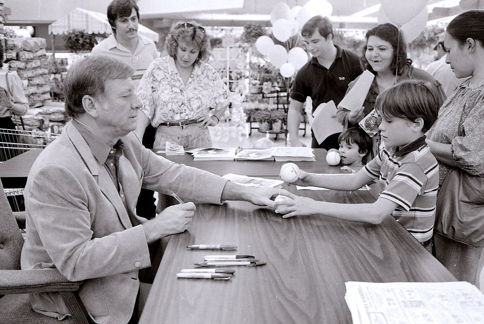 A black and white photograph of Mickey Mantle signing baseballs for 9-year-old Jeremy Caird at Dillons, a grocery store. Other fans look on.