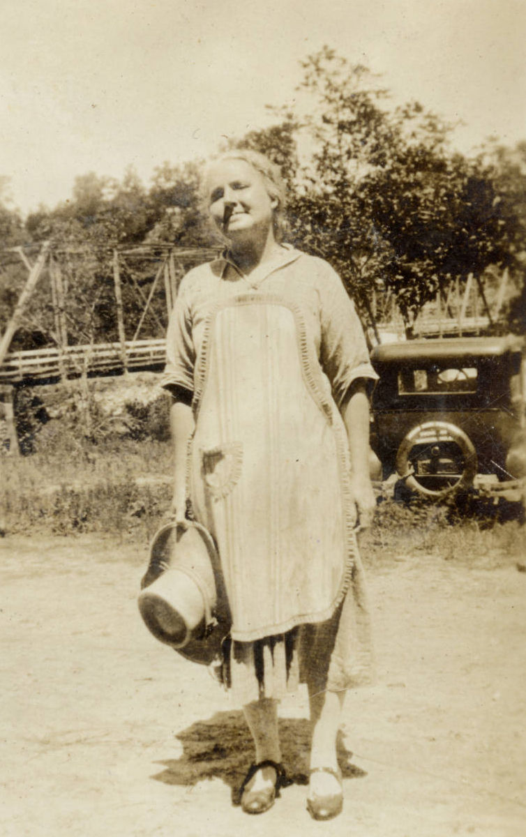 Full length portrait of Mary Elizabeth Mahnkey taken in the 1930s. Mahnkey is wearing a dress, apron, and is holding a hat.