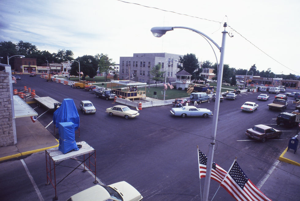 An elevated view of the Marshfield square during preparations for the 1991 Fourth of July parade. A reviewing stand and a stage are in the interior portion of the square, near the courthouse. Traffic moves around the square.
