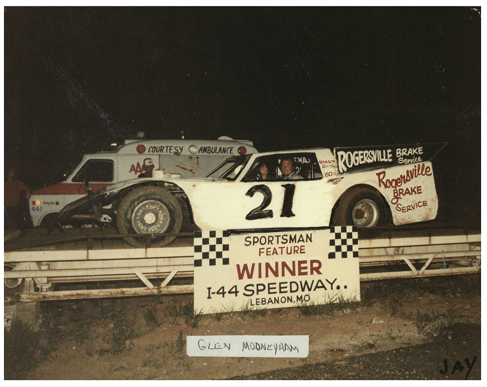 Driver Glen Mooneyham sits in the drivers seat of his race car with a sign that reads "Sportsman Feature Winner I-44 Speedway. Lebanon, Mo." The livery on Mooneyham's car reads "Rogersville Brake Service."