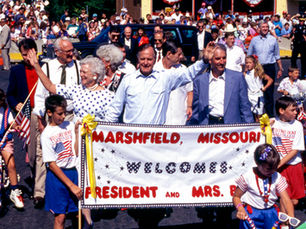 President George H. W. Bush and First Lady Barbara Bush lead the Fourth of July parade in Marshfield, 1991. Many people fill the frame. A sign 