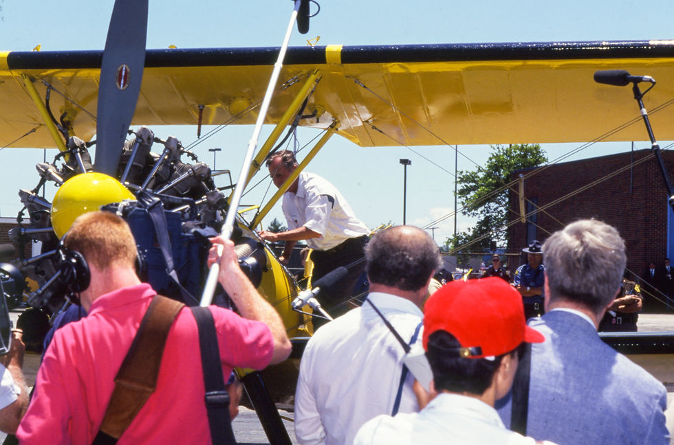 President George H. W. Bush stands on the wing of a yellow Stearman biplane and peers into its cockpit while journalists look on.
