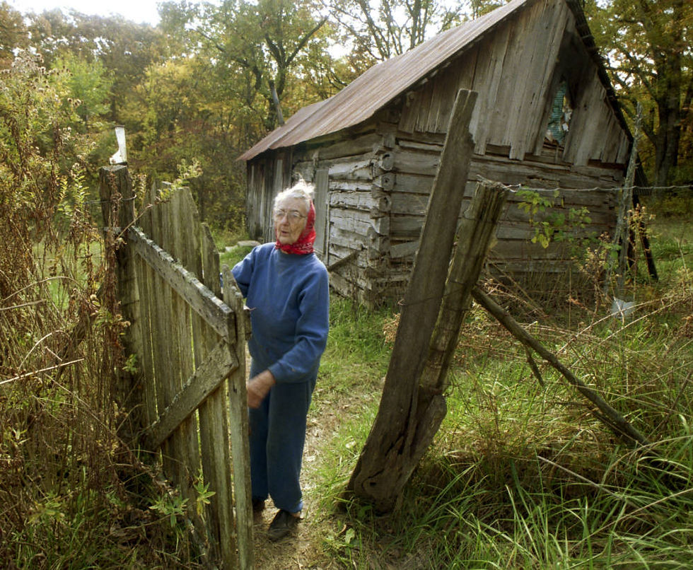 A photograph of an elderly woman, Birdle Mannon, closing the gate in front of her rustic Ozarks cabin.