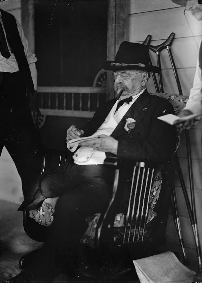 An old man sitting in a rocking chair and wearing a felt hat, glasses, and a suit dangles a cigar from his mouth as he signs a paper. His right pantleg is folded at the thigh.